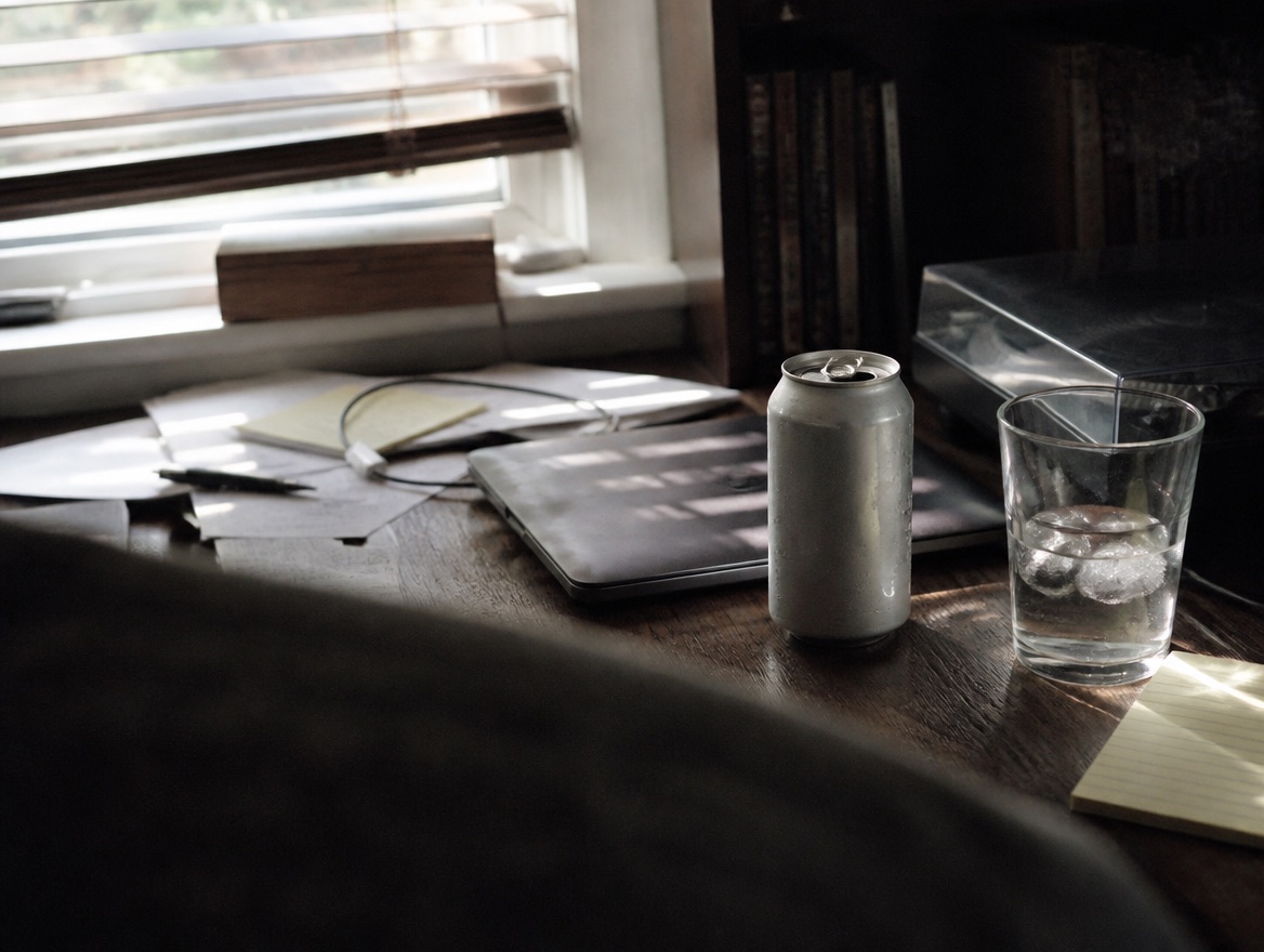 An open, condensation-beaded soda can sits on a cluttered wooden desk beside a closed laptop and a glass of ice water, with papers, a notebook, and a phone charger cord in afternoon light from a window blind.