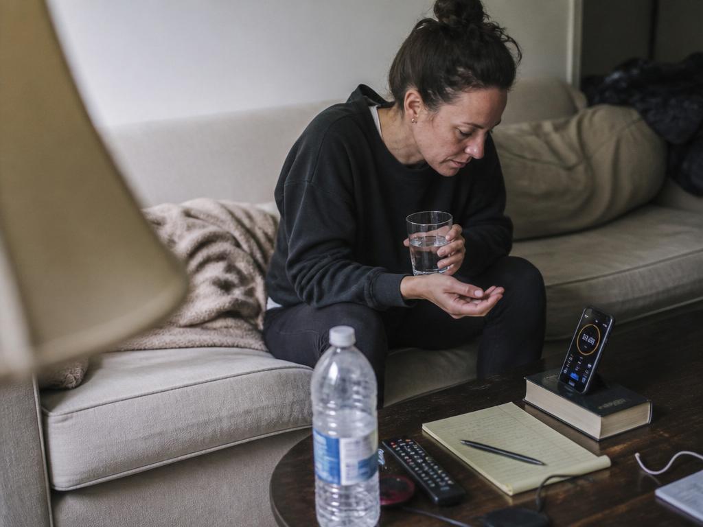 A woman on a long fast sits on a couch looking depleted, holding a glass of water beside a phone showing a timer.