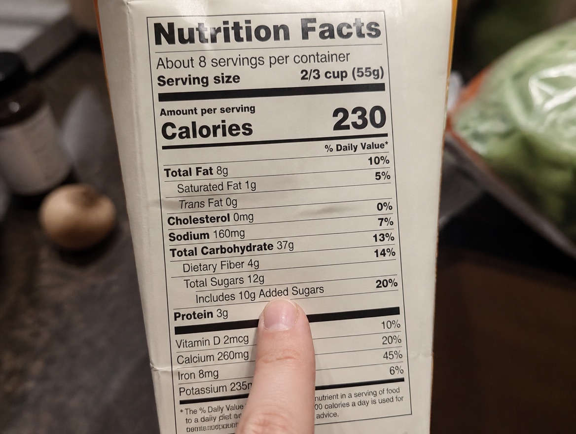 An extreme close-up of a nutrition facts panel on a food box, held under bright kitchen light, with a fingertip resting near the added sugars line below total carbohydrate and dietary fiber, a blurred kitchen counter behind it.